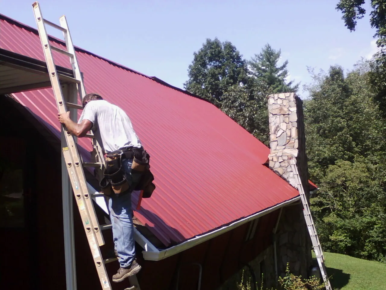Roofer on a ladder beside a red metal roof and stone chimney in Avon